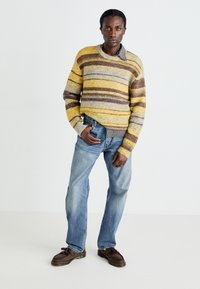 Suéter de rayas en amarillo, gris y marrón; puños acanalados; llevado sobre una camisa de cuadros con jeans azules y mocasines marrones.