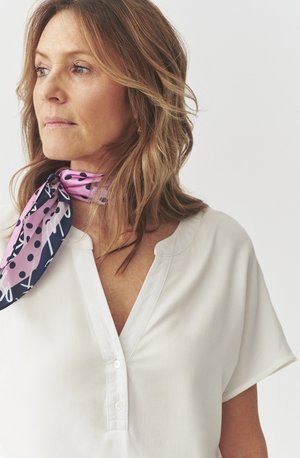 Blusa blanca con un diseño abotonado y un leve escote en V. Una bufanda rosa y azul marino con lunares está atada alrededor del cuello, añadiendo un acento contrastante.