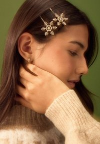 Guldtonade hårnålar formade som snöflingor med klara rhinestones, satta i mörkbrunt hår, mot en grön bakgrund; mjuk beige tröja synlig.