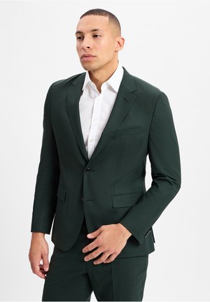 Donkergroene pakjas met een enkele knoop, gelapte revers en twee voorzakken. Heeft een witte overhemd eronder.
