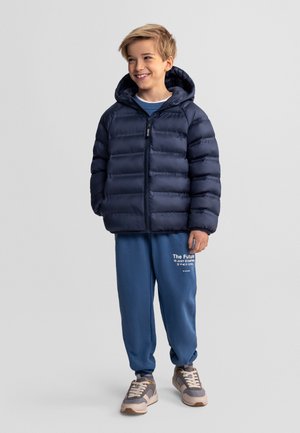 Usměvavý chlapec v tmavě modré prošívané bundě s kapucí, modrých teplácích s bílým nápisem a šedých teniskách stojí na jednobarevném šedém pozadí.