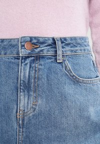 Ljust blå denimkjol med hög midja, prydd med kopparfästen, sidofickor och en diskret urtvättad textur.