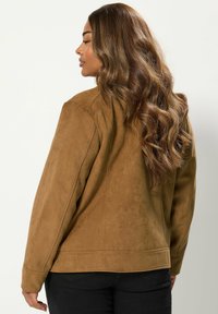 Kvinna med långt, vågigt brunt hår som bär en brun mockajacka och svarta byxor, vänd åt sidan mot en enkel ljus bakgrund.