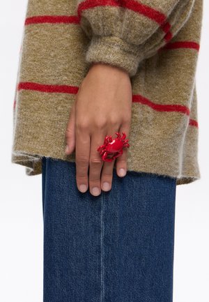 Anillo en forma de cangrejo rojo en una mano, presentado con un suéter de punto verde-marrón texturizado y denim, con rayas rojas.
