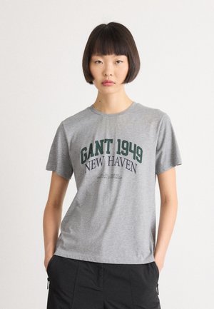 Young woman with short dark hair wearing a gray GANT 1949 New Haven t-shirt and black pants with hands in pockets.