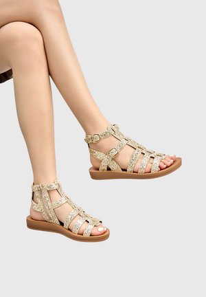 Jambes portant des sandales plates dorées pailletées à brides avec boucles, sur un fond gris clair uni.