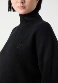 Femme portant un pull noir à col montant avec un petit logo smiley noir brodé sur la poitrine.