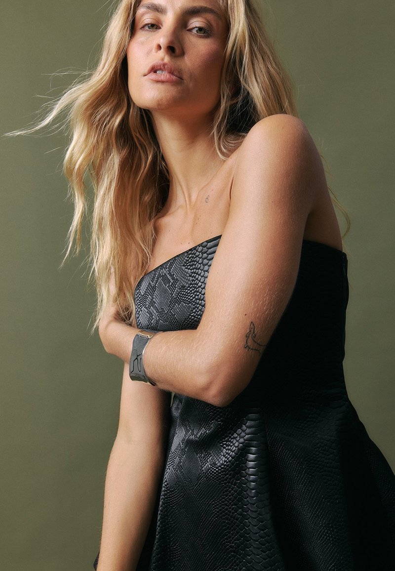 Juodas be petnešų suknelė su tekstūruotu gyvatės odos raštu, turinti prigludusį korsetinį viršų ir platų sijoną. Sidabrinė apyrankė ant modelio rankos.