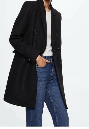 Person iført en sort dobbeltradet frakke over en hvid skjorte med blå jeans, med den ene hånd i forlommerne på jeansene.