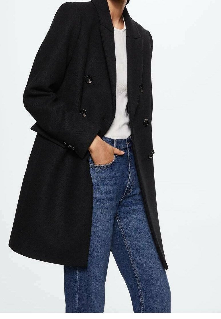 Personne portant un manteau noir croisé sur une chemise blanche avec un jean bleu, une main dans la poche avant du jean.