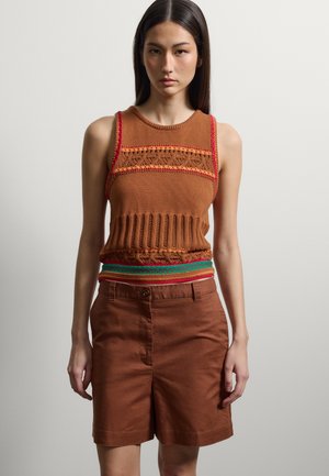 Junge Frau mit langen dunklen Haaren, die ein ärmelloses braunes Strickoberteil mit rotem und grünem Besatz trägt, kombiniert mit braunen hoch taillierten Shorts.