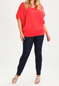 Top rosso ample con scollatura a V e fondo elasticizzato, abbinato a jeans skinny blu scuro e sandali chiari con ricami floreali.