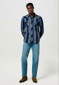 Marinblå langermet skjorte med vertikale striper i lysere nyanser, to brystlommer og snap-knapper, sammen med lyseblå jeans og brune støvler.