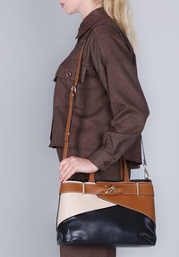 Vrouw in een bruine outfit die een grote leren handtas vasthoudt met zwarte, beige en bruine kleurvakken en een metalen sluiting.