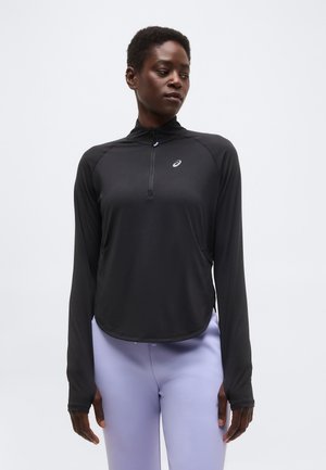 Žena oblečená v čiernom športovom tričku s dlhým rukávom a polovičným zipsom a v svetlofialových legínach stojaca pred jednoduchým svetlým pozadím.