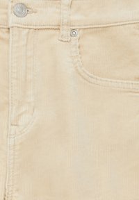 Pantalon en velours côtelé beige en gros plan montrant la poche avant, le rivet métallique et les détails de la braguette à boutons.
