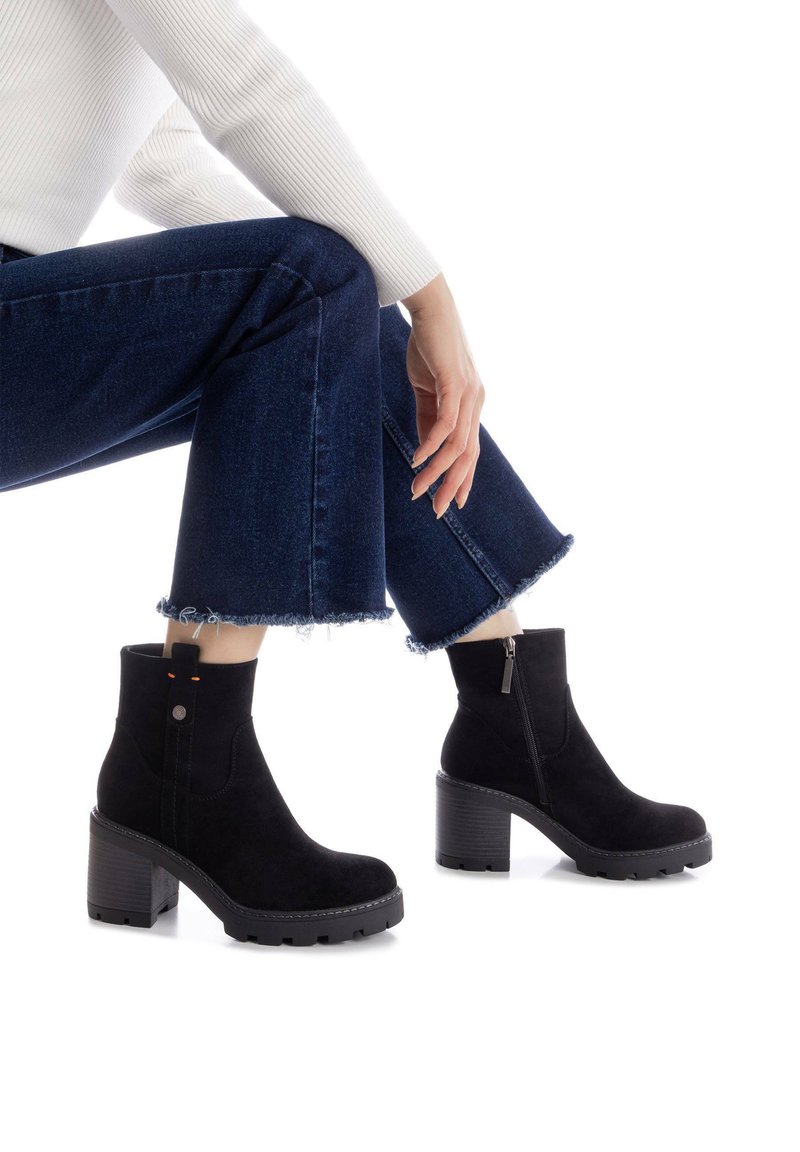 Botas de ante negras hasta el tobillo con un tacón grueso, cremallera lateral y costuras en contraste. Llevadas con jeans oscuros de pierna acampanada con puños desgastados.