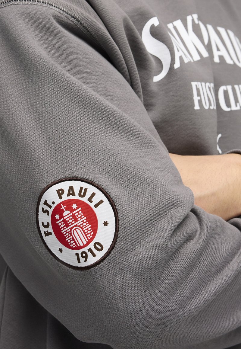 Sudadera gris con una textura suave. Presenta un logo redondo del FC St. Pauli en la manga izquierda, con colores rojo, blanco y marrón.