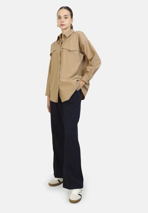 Tan button-up shirt with a pointed collar, chest pockets, and side slits, paired with high-waisted navy trousers and white sneakers.