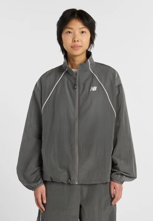 Nuori henkilö pukeutunut harmaaseen New Balancen vetoketjulliseen urheilutakkiin ja sopiviin housuihin, seisoo yksinkertaisen vaalean taustan edessä.