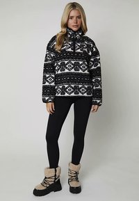 Svartvit mönstrad fleece-tröja med hög krage, matchad med svarta leggings och bruna pälsfodrade stövlar.