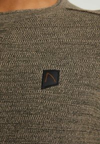 Olivgrüner Pullover mit strukturiertem Strickmuster. Mit einem schwarzen Patch mit einem orangefarbenen Logo auf der linken Seite nahe der Schulter.