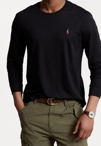 Zwart T-shirt met lange mouwen en ronde hals, met geborduurd logo; gedragen met olijfgroene cargobroek en een bruine riem, voorzien van een polshorloge.