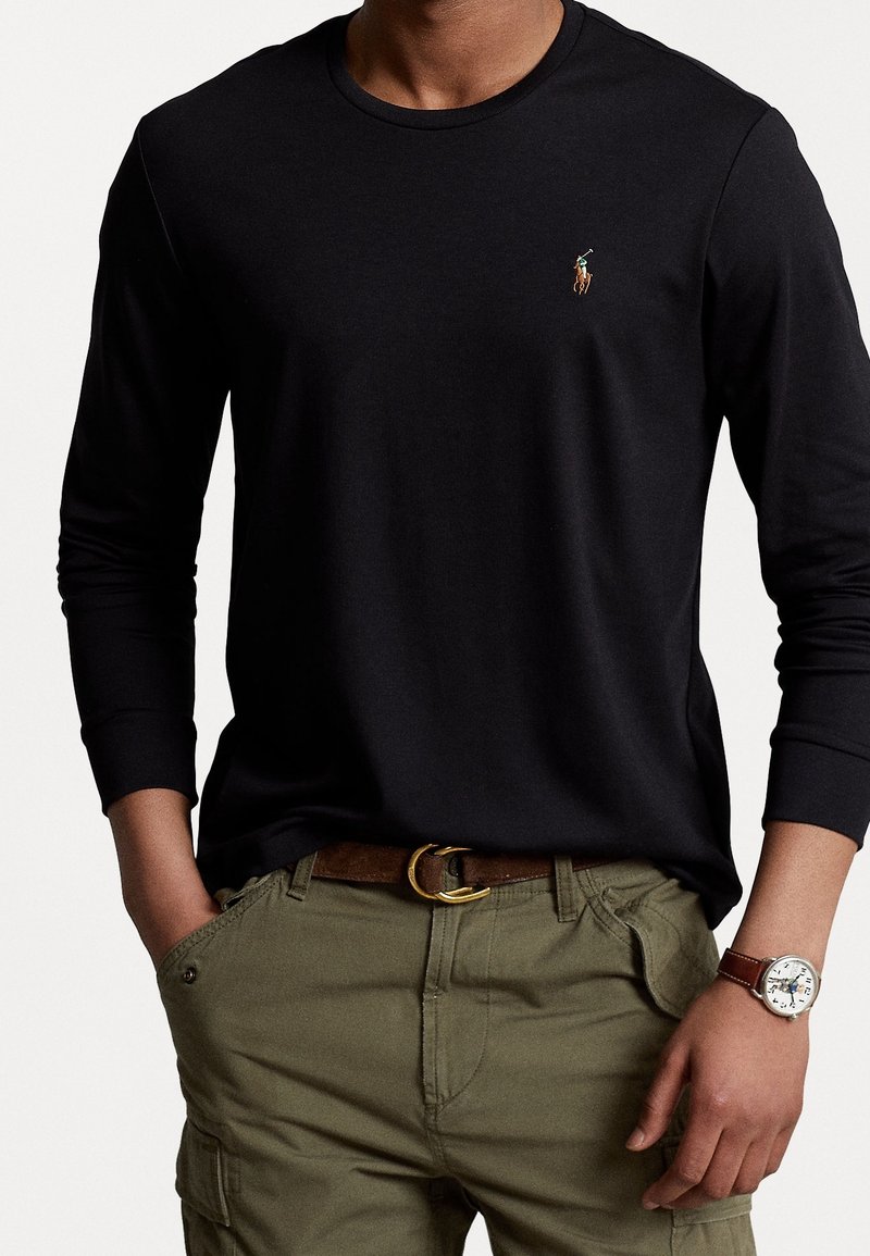 Zwart T-shirt met lange mouwen en ronde hals, met geborduurd logo; gedragen met olijfgroene cargobroek en een bruine riem, voorzien van een polshorloge.