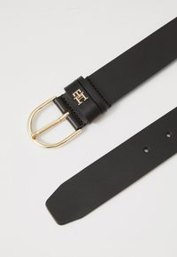 Ceinture en cuir noir avec une boucle rectangulaire en or. Présente un emblème de logo sur le passant en cuir. Texture lisse avec des trous régulièrement espacés.