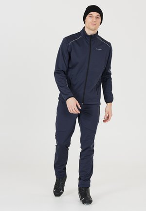 Veste de sport et pantalon bleu marine en tissu extensible, avec fermeture éclair à l'avant, poches latérales et coupe ajustée. Bonnet noir sur la tête.