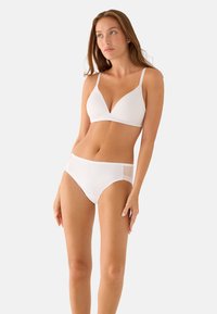 Mujer con sujetador blanco sin costuras y ropa interior de talle alto a juego, de pie con postura relajada contra un fondo liso y claro.