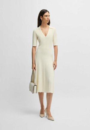 Abito a coste, lungo fino al ginocchio, di un crema chiaro con scollo a V e maniche corte. Abbinato a una borsa leggera e tacchi beige affusolati.
