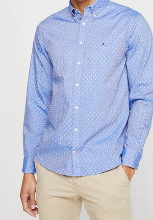 Mand iført en lyseblå skjorte med hvide polkaprikker og beige bukser, den ene hånd i lommen.