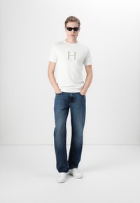 Hvit t-skjorte med en grønn "H"-logo, kombinert med mørkeblå jeans og hvite sneakers. Modell iført svarte solbriller, står med hendene i lommene.