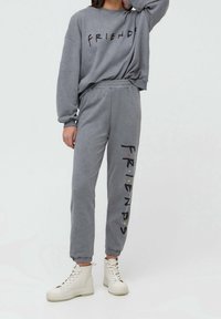 Person iført grå loungedragt med "FRIENDS" tekst på sweatshirt og bukser, parret med off-white højtop sneakers, stående mod hvid baggrund.