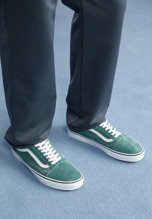 Osoba nosząca ciemne spodnie i turkusowo-zielone trampki z białymi paskami i sznurówkami stojąca na jasnoniebieskim wykładzinowym podłodze.