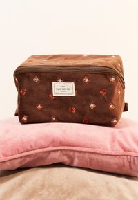 Ruskea kangaslaukku, jossa on kukkakirjontaa, vetoketjukiinnitys ja etiketti. Asetettu pinkkien ja beigekuiden pehmeiden tyynyjen päälle. Suede-tyylinen pinta.