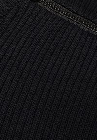 Czarna prążkowana tkanina z widocznym detalem zamka błyskawicznego. Faktura jest dzianinowa, zapewnia dopasowanie i gładki wygląd.