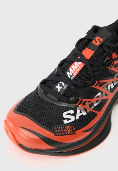Černá a oranžová sportovní bota s síťovaným svrškem, výrazným logem, texturovanými detaily a výraznou zakřivenou podrážkou pro podporu.