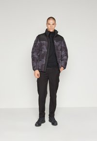 Giacca puffa in camo scuro con una sezione superiore nera, abbinata a un giubbino nero con zip e pantaloni cargo neri. Calzature nere.