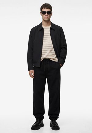 Mężczyzna w czarnych okularach przeciwsłonecznych, czarnej kurtce, beżowej koszuli w paski, czarnych spodniach dresowych i czarnych masywnych butach, stojący z jedną ręką w kieszeni.