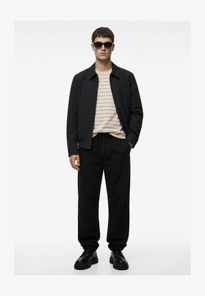 Mężczyzna w czarnych okularach przeciwsłonecznych, czarnej kurtce, beżowej koszuli w paski, czarnych spodniach dresowych i czarnych masywnych butach, stojący z jedną ręką w kieszeni.