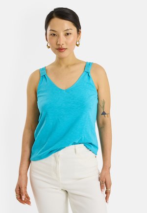 Mujer con cabello oscuro, pendientes de aro dorados, top sin mangas turquesa, pantalones blancos y un tatuaje en su brazo izquierdo, de pie frente a un fondo blanco.