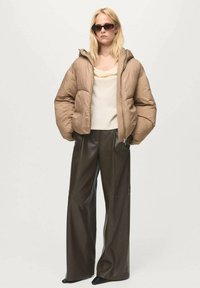 Giubbotto a vento di colore marrone chiaro con cappuccio, abbinato a un top crema e pantaloni a palazzo in pelle sintetica marrone scuro. Il modello indossa occhiali da sole scuri.