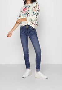 Blommönstrad ljus sweatshirt, blå skinny jeans och vita sneakers. Sweatshirten har långa ärmar och ribbstickade muddar; jeansen har framfickor.