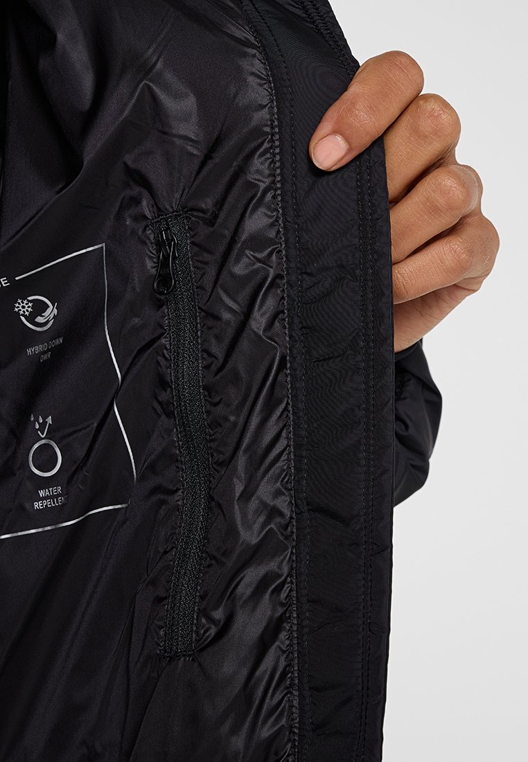 Eine Hand hält eine offene schwarze Jacke, die eine innere Reißverschlusstasche und Stoffetiketten zeigt, die auf Hybrid-Daunenisolierung und Wasserabweisbarkeit hinweisen.