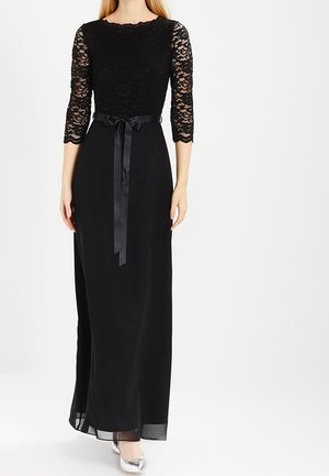 Kobieta w długiej czarnej sukni z koronkowymi rękawami do połowy przedramienia i satynowym paskiem oraz srebrnymi szpilkami.