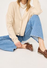 Femme assise avec un genou relevé, portant un cardigan beige, une blouse blanche, un jean large bleu clair et des mocassins en cuir marron sur un sol blanc.