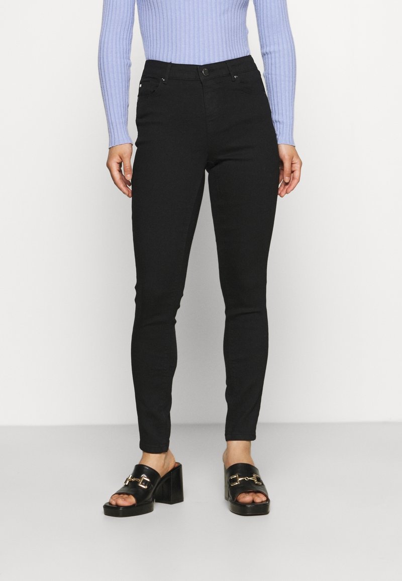 Jeans negros ajustados con una cintura alta, que cuentan con dos bolsillos frontales, un cierre de botón y una textura suave, combinados con unas sandalias negras de tacón.