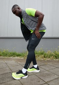 Conjunto atlético composto por um colete acolchoado cinza, uma camiseta de manga curta verde limão, leggings pretas e tênis de corrida com detalhes verdes e solas brancas.